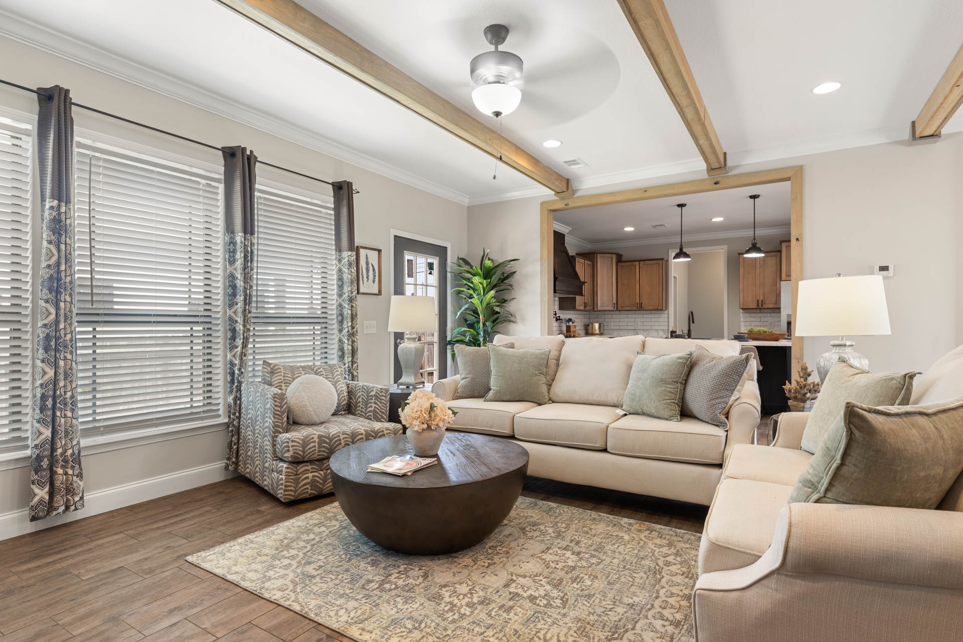 Modern living room with beige sofas, patterned pillows, and a round coffee table on a patterned rug. Large windows with drapes and a ceiling fan add brightness.