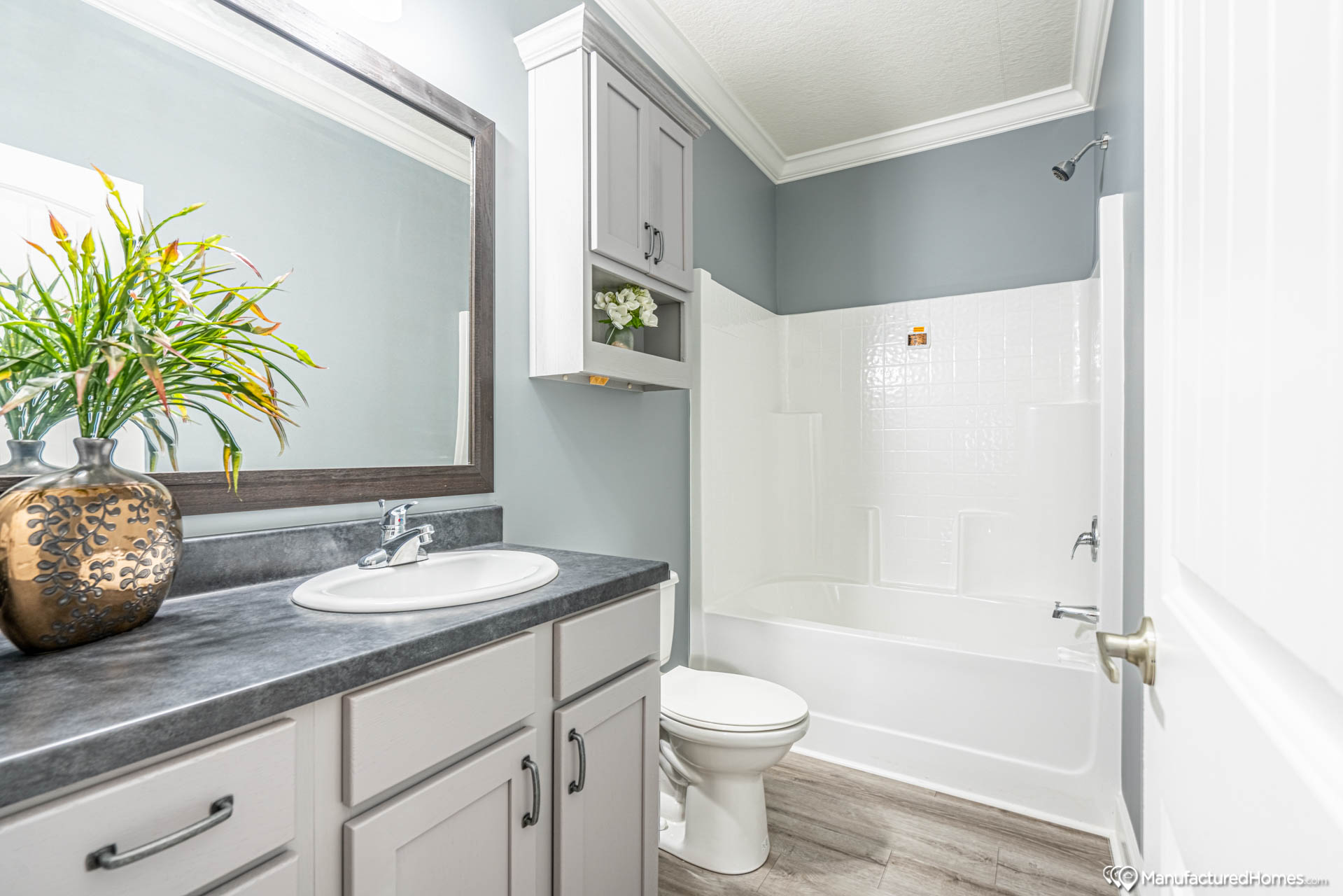 A modern bathroom with light gray walls. It features a vanity with a dark countertop, round sink, and an ornate vase with greenery. A white tub, gray cabinets, and a serene, clean atmosphere complete the space.