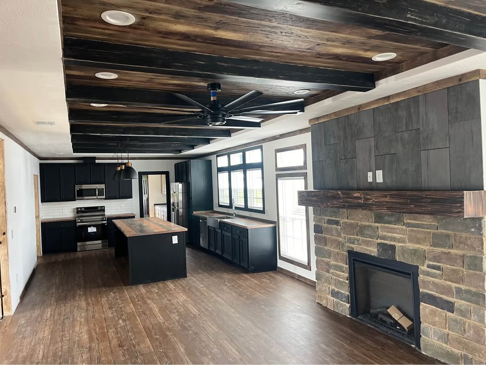 Spacious kitchen and living area with dark hardwood floors, a large ceiling fan, stone fireplace, and modern black cabinetry under a beamed ceiling.