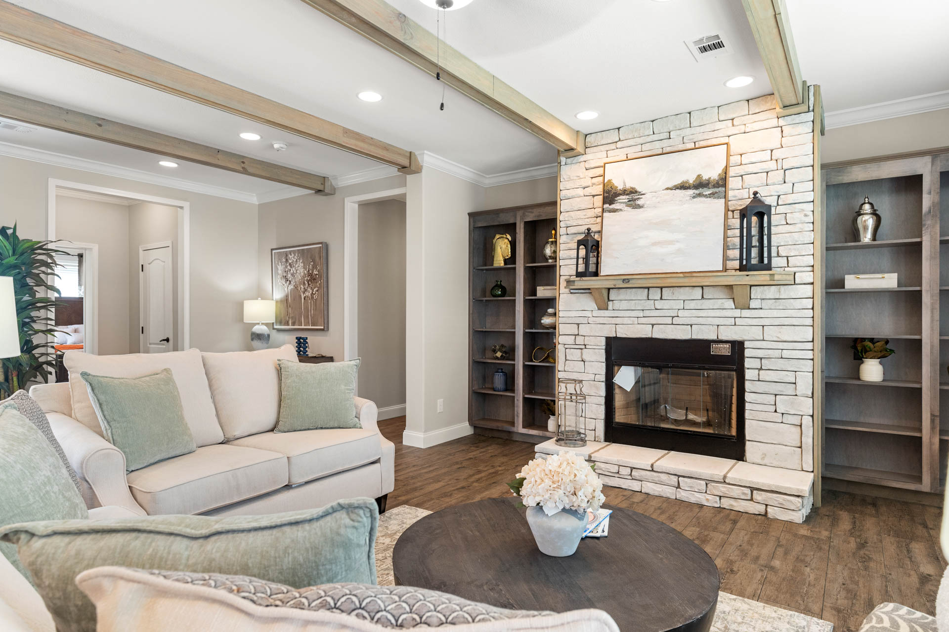 Cozy living room with a white sofa, pastel cushions, and a wooden coffee table. Stone fireplace with artwork, shelves, and warm lighting create a welcoming ambiance.