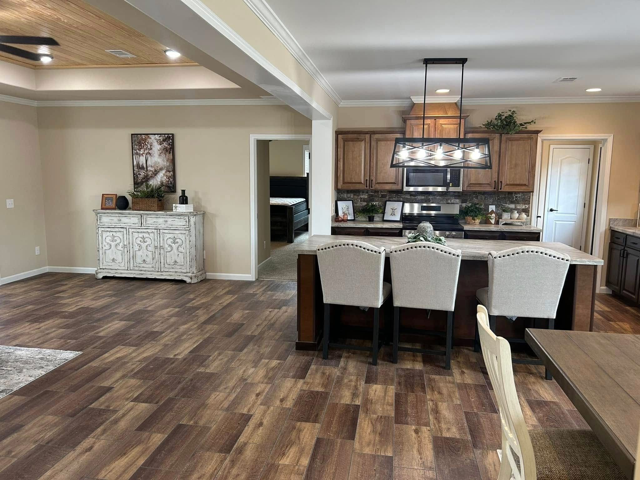 A modern kitchen and dining area with wood flooring, a central island with light grey chairs, pendant lighting, and wooden cabinets conveying warmth.