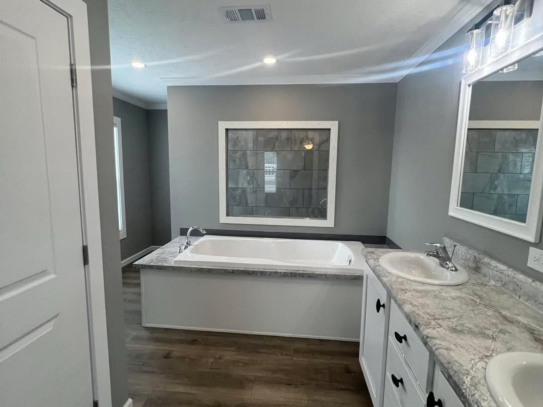 Modern bathroom with a large bathtub set against a gray tiled wall. Marble countertop with twin sinks and mirrors, creating a clean, serene atmosphere.