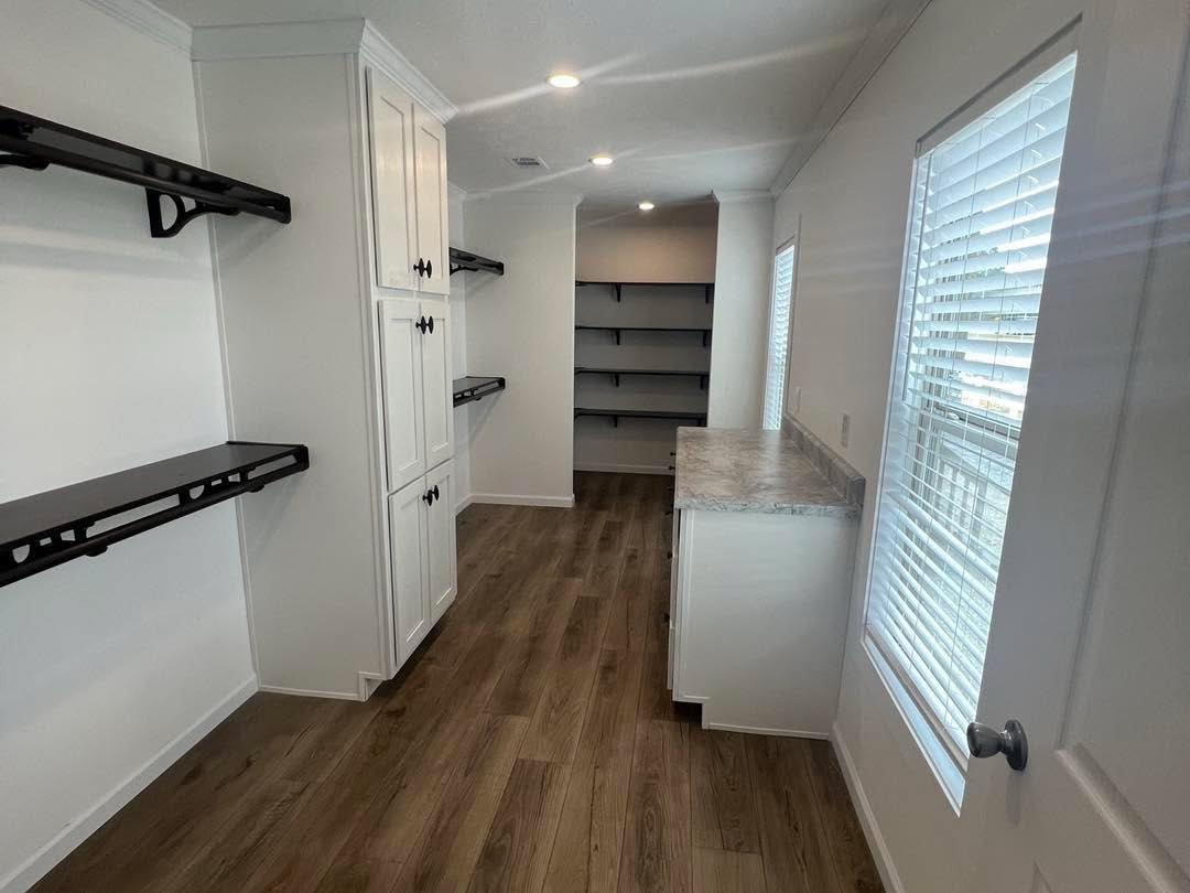 Bright pantry with wood floors, white walls, black shelves, and cabinets. A long countertop is on the right, under a window with blinds. The mood is clean and organized.