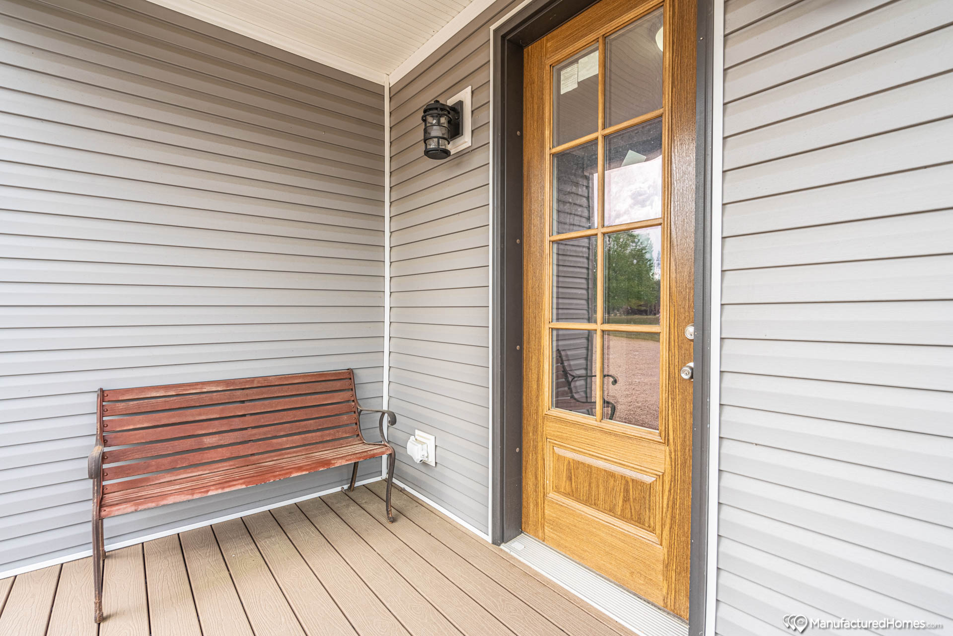 A wooden bench and a glass-paneled wooden door are on a porch with gray siding. A black lantern is mounted above, creating a welcoming atmosphere.