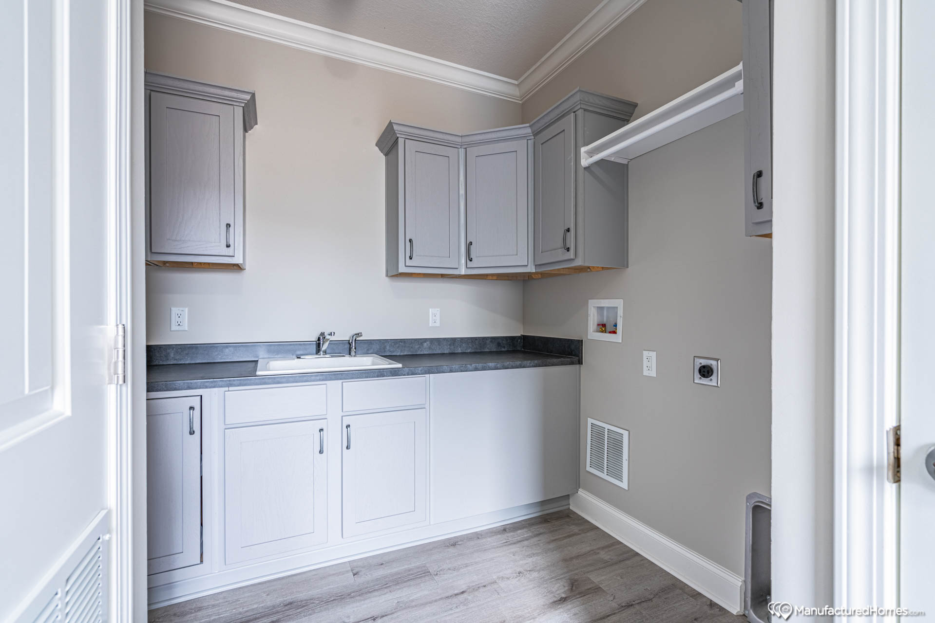 Laundry room with light gray cabinets, countertop, and sink against a beige wall. Wood flooring adds warmth. Space for washer and dryer noted.