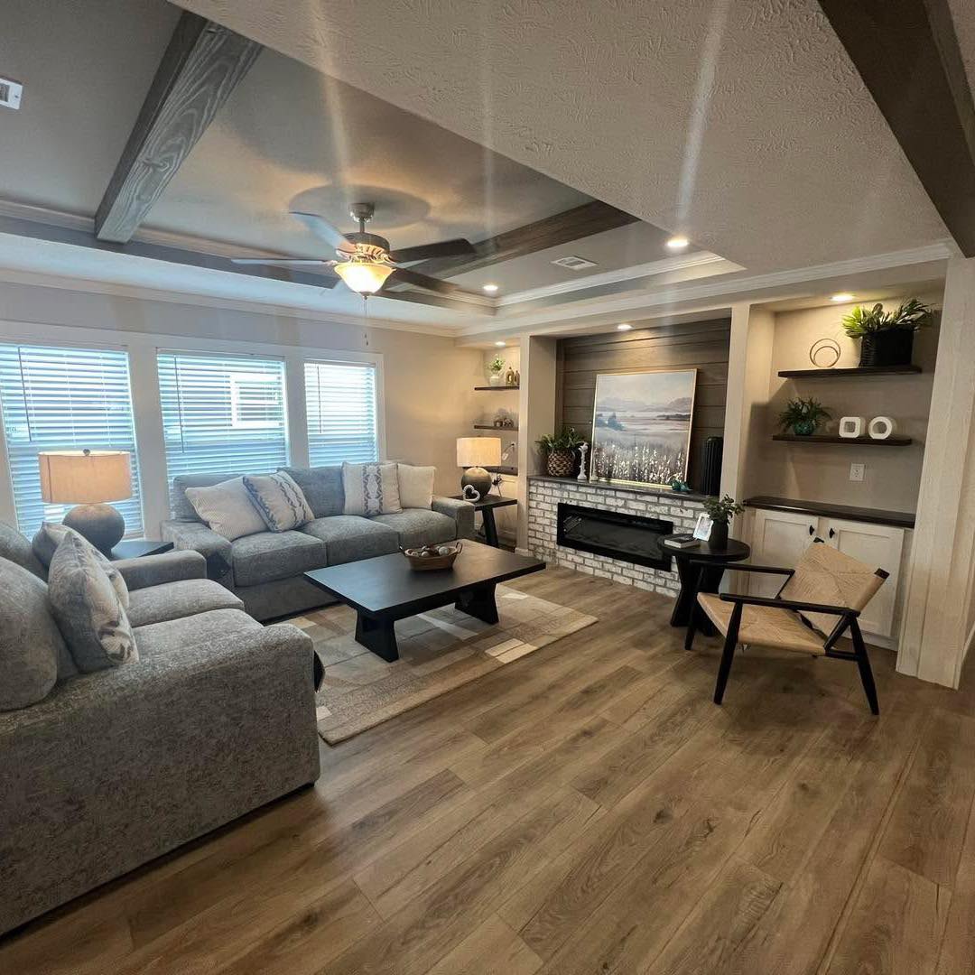 Spacious living room with a cozy gray sectional, wooden coffee table, and beige armchair. Ceiling beams enhance the modern decor. A lit fireplace and artwork add warmth.