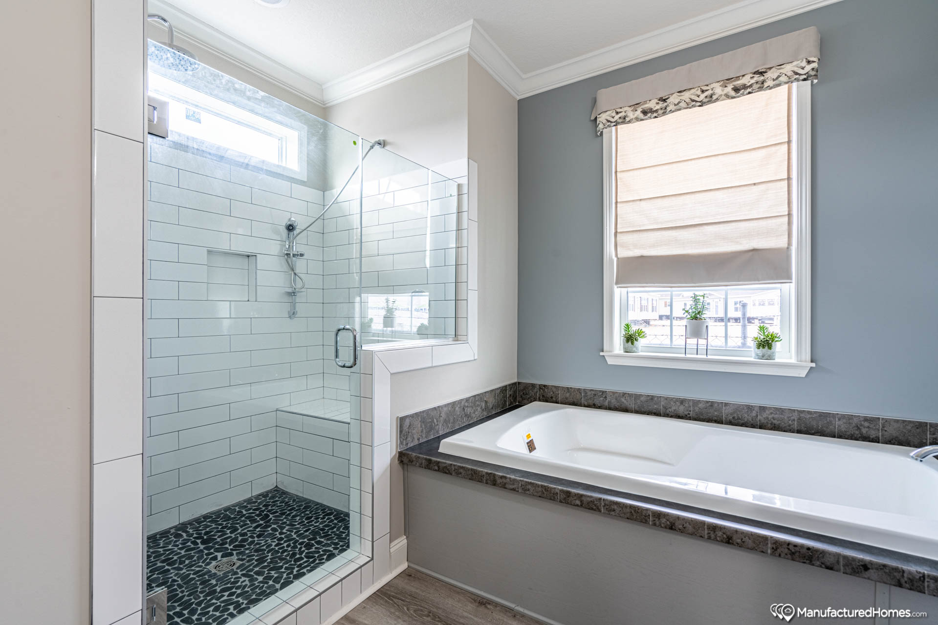 Modern bathroom with glass-enclosed shower featuring dark pebble tile floor, light tiled walls, adjacent to a white bathtub with gray countertop, under a window with a beige shade.