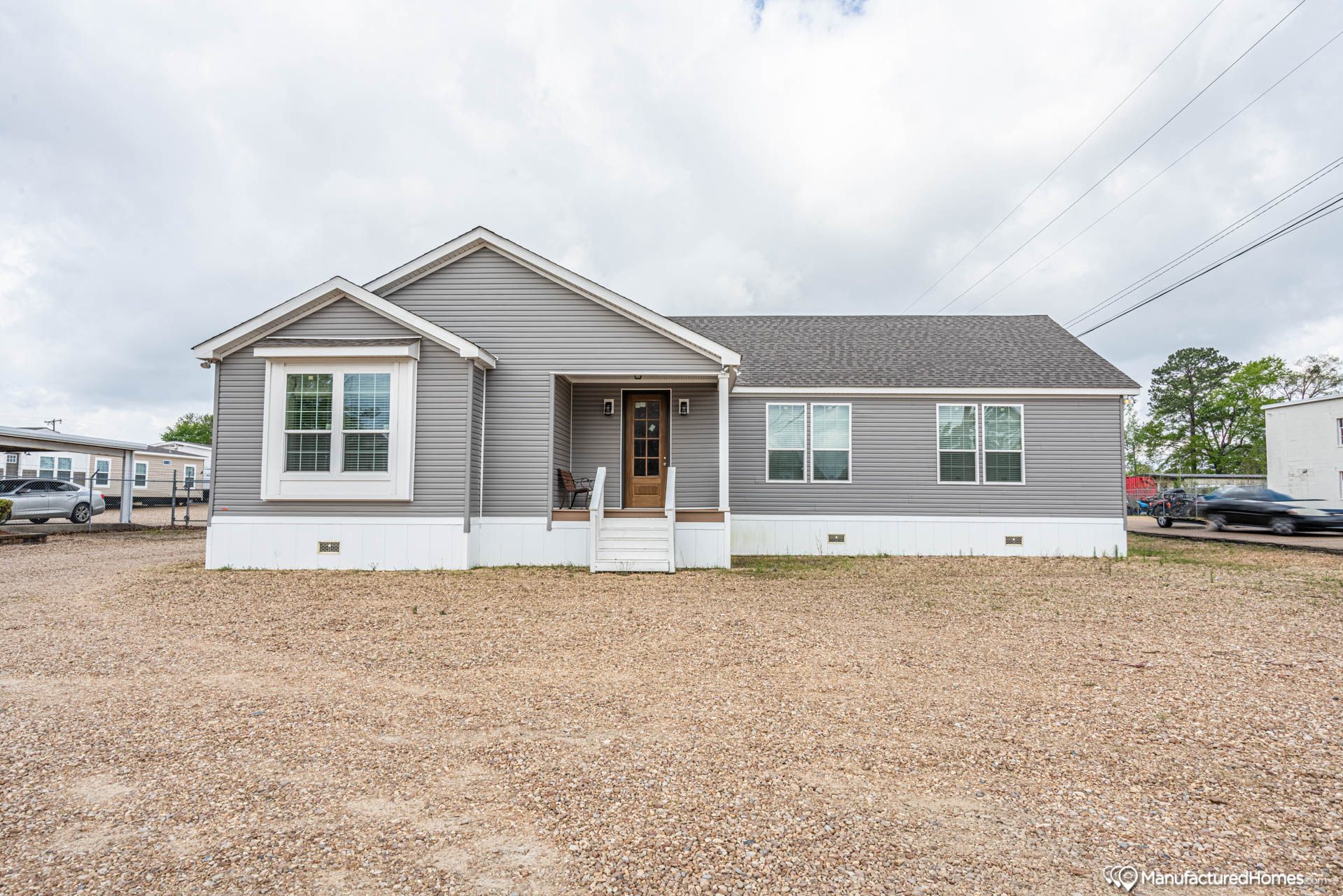 Single-story gray manufactured home with a gable roof and white trim, set on a gravel lot. Small porch with a chair, overcast sky overhead.