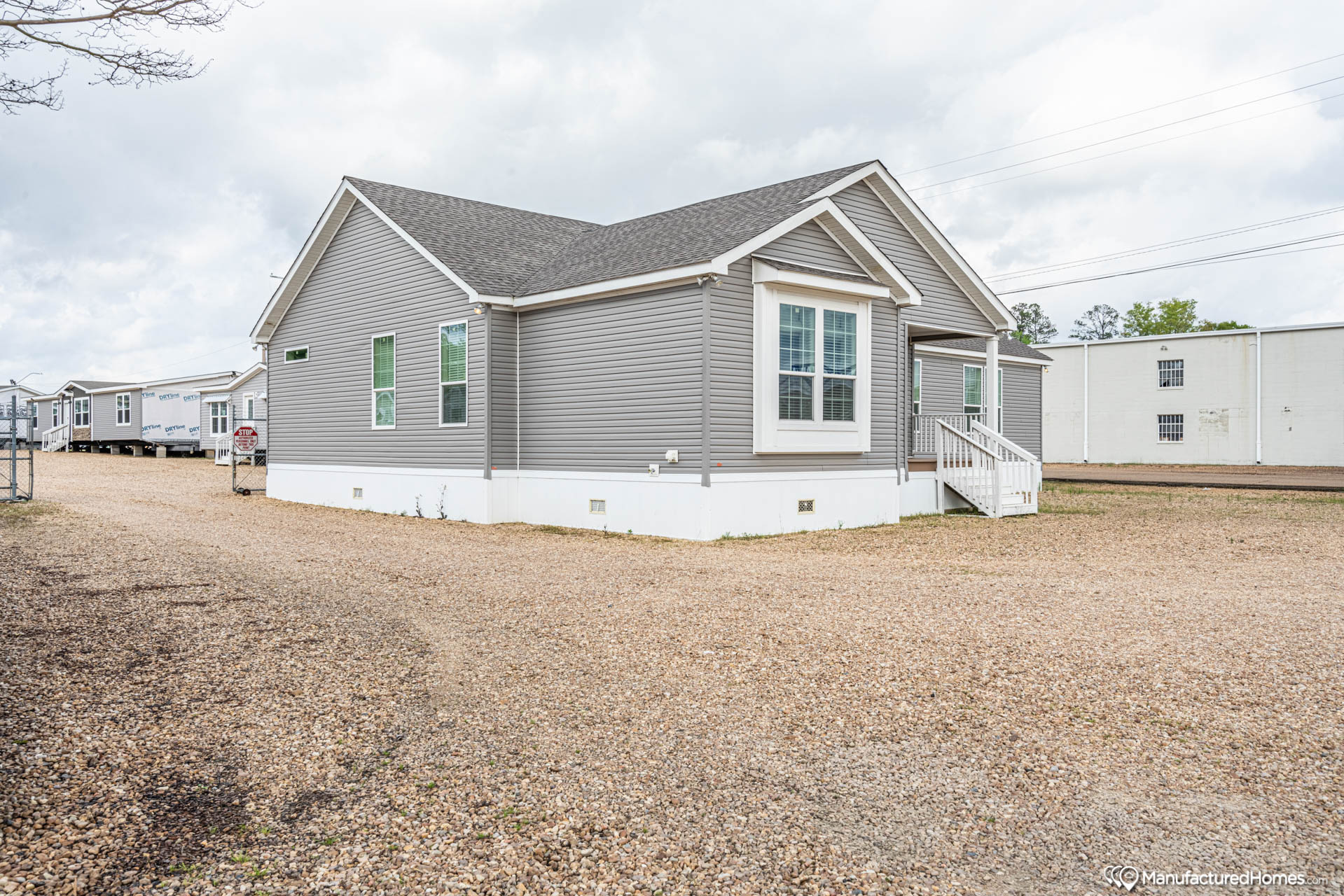 A gray manufactured home with white trim is set on a gravel lot under a cloudy sky. The home features multiple windows and a small porch with steps.