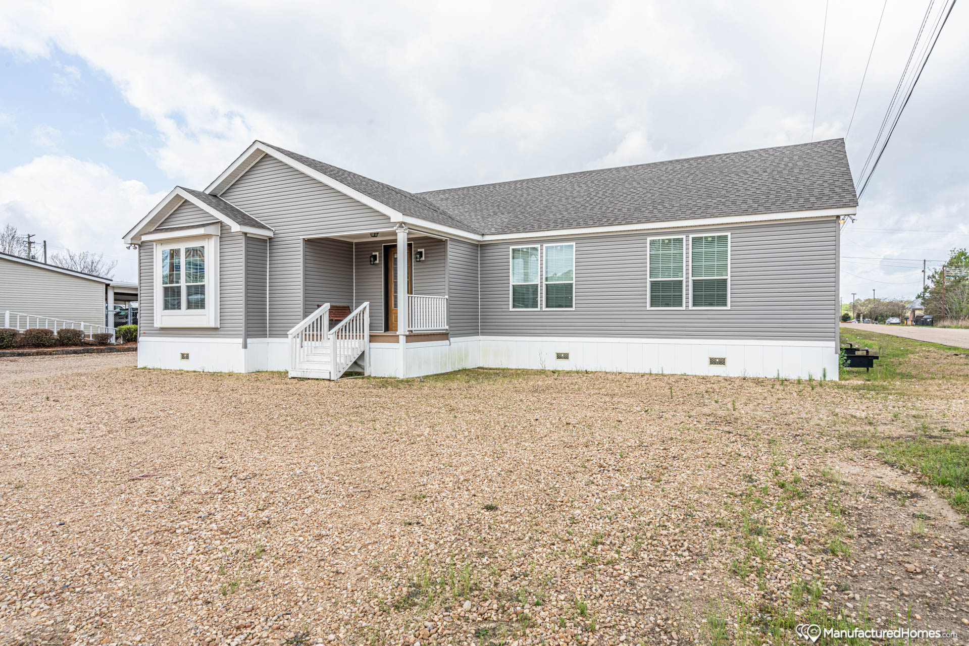 A single-story gray manufactured home with white trim sits on a gravel lot. It has a small porch with stairs, large windows, and a cloudy sky above.