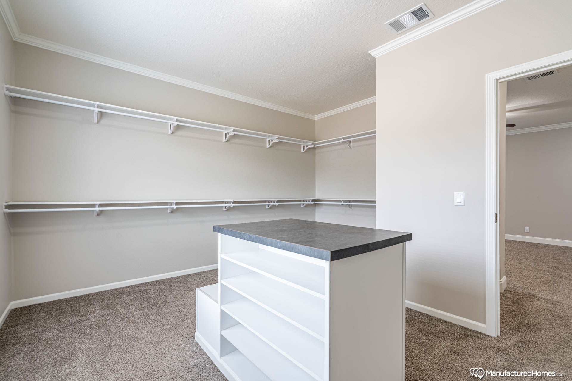 Spacious walk-in closet with beige walls and carpet, featuring white shelves and hanging rods. A central island has open shelving and a dark countertop.