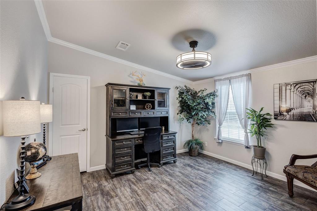 Spacious home office with dark wooden desk, black chair, and a hutch against a light gray wall. Large potted plants and sheer-curtained windows add warmth.