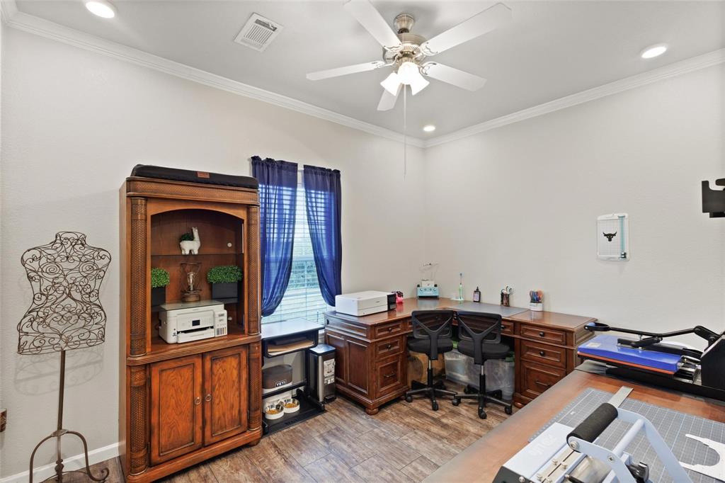 Home office with wooden flooring, featuring a ceiling fan, vintage wooden desk, and shelves. Blue curtains adorn the window, and a mannequin form stands nearby. The room is neatly organized, conveying a warm, professional atmosphere.
