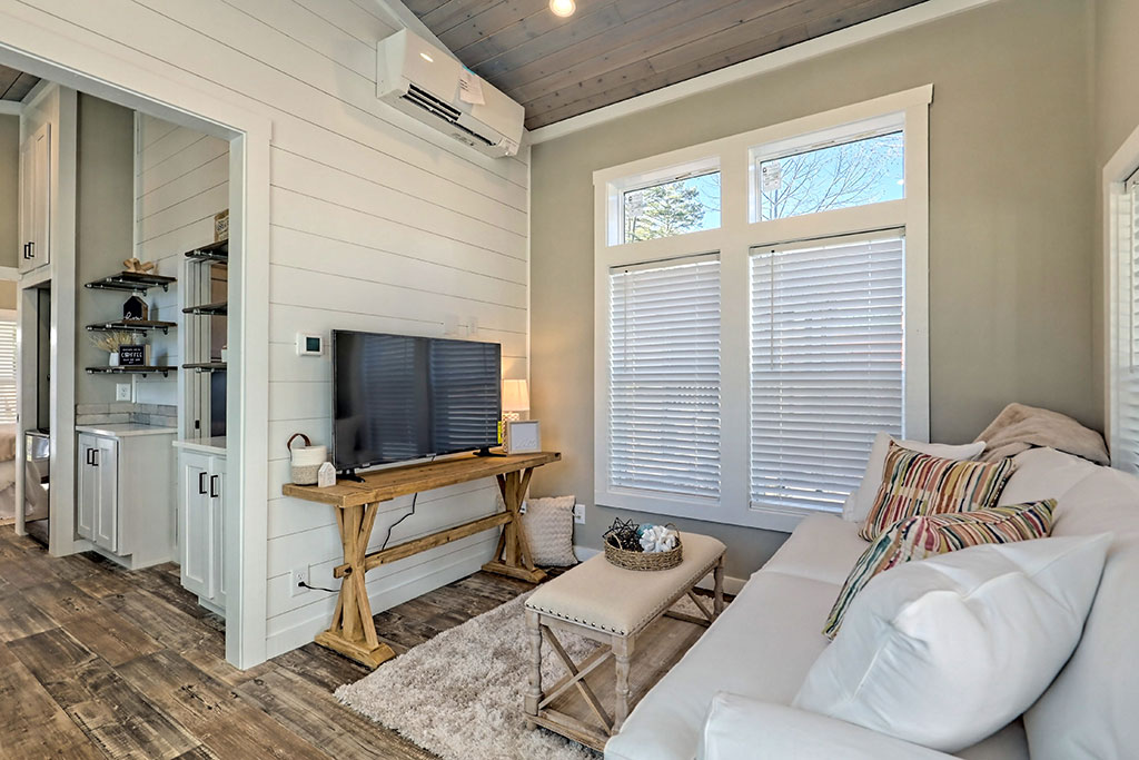 Cozy living room with a white couch and colorful pillows. A wooden table holds a TV beneath windows with blinds. Rustic wood flooring and minimalist decor.