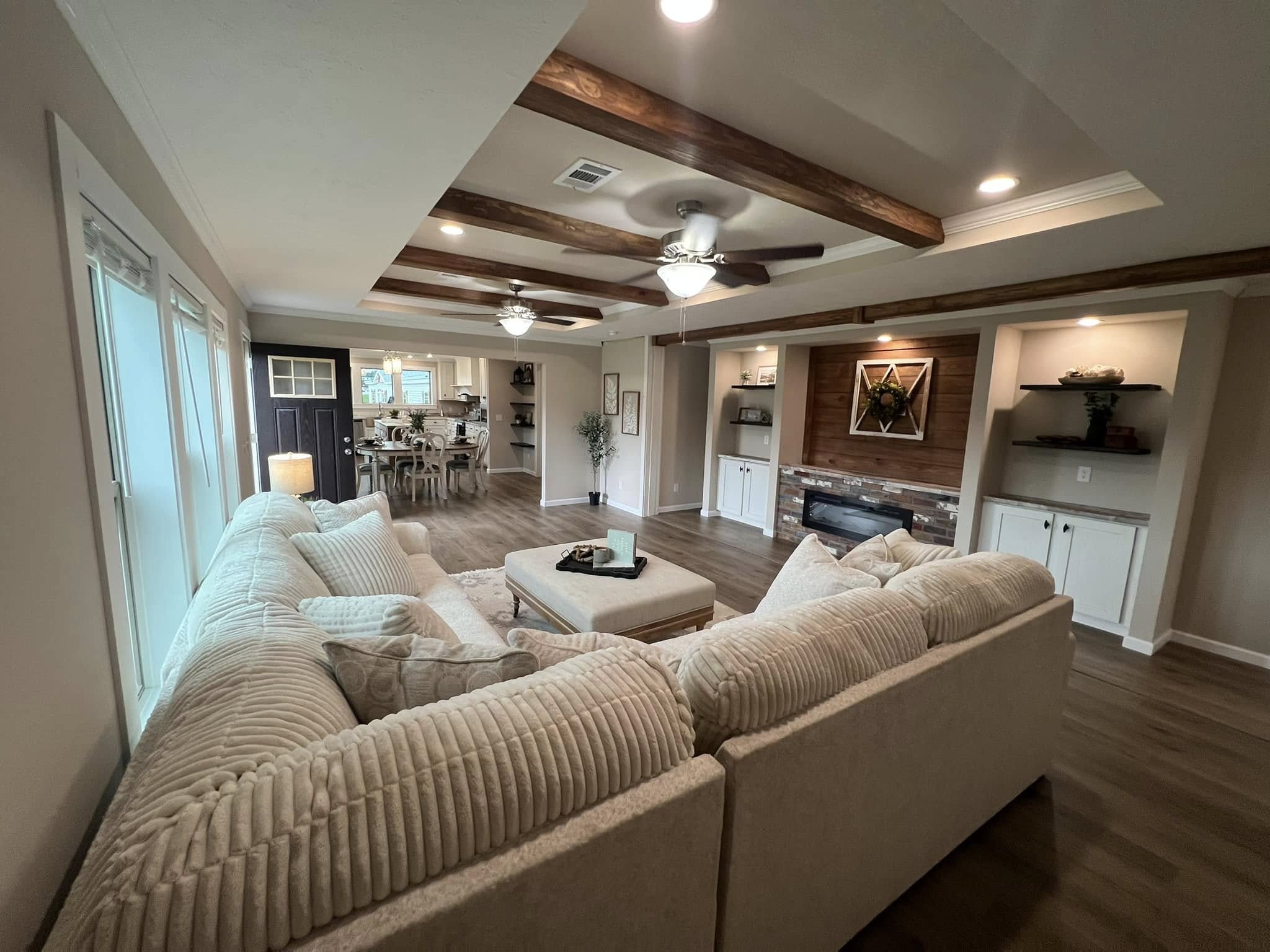 Spacious living room with beige sectional sofa, wooden beams on ceiling, light wood floor, and a fireplace. Dining area visible in background. Cozy and inviting ambiance.