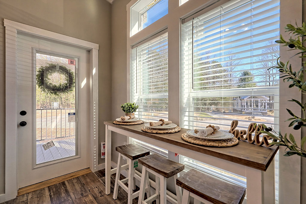 Cozy sunlit dining corner with a rustic wooden table set for four, framed by tall windows with blinds. A wreath hangs on the door, creating a welcoming vibe.
