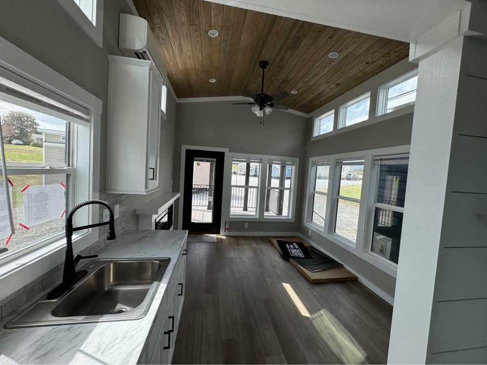 A bright, modern tiny house interior with wood floors and ceiling. The room features large windows, a ceiling fan, and a compact kitchen with a marble countertop.