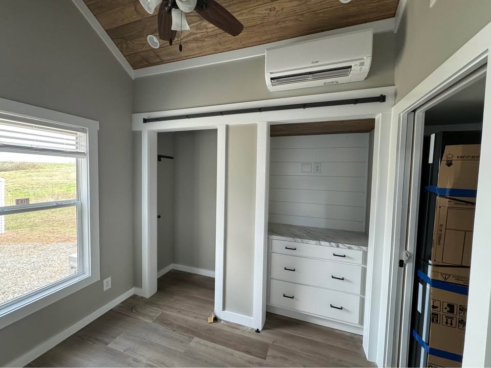 A cozy room with a wooden ceiling and light gray walls, featuring an open closet with sliding doors, drawers, and an air conditioner above. A window allows natural light.