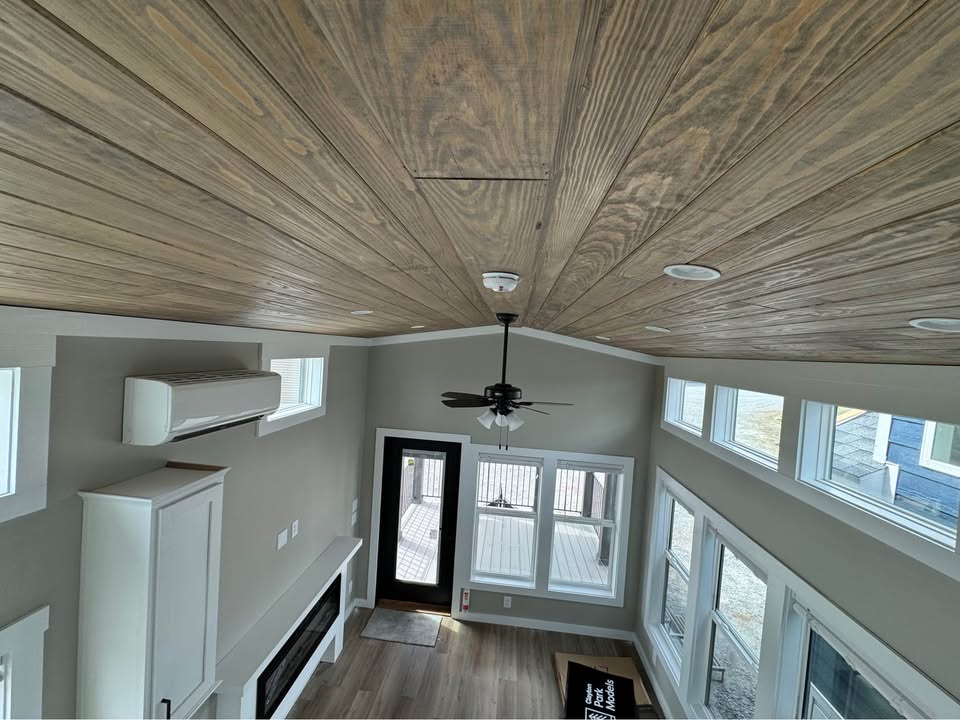 High-angle view of a modern tiny house interior. Features include wide wooden ceiling, ceiling fan, large windows, light wood flooring, and a small fireplace, conveying a cozy, airy atmosphere.