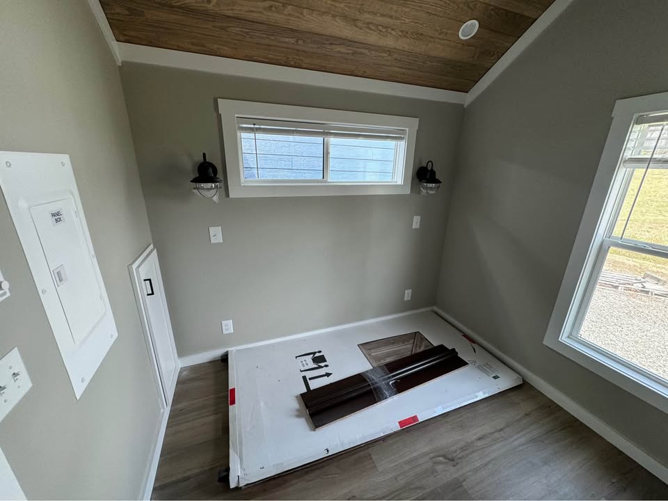 Small room under renovation with wooden floor, gray walls, and a wood-paneled ceiling. Two wall lamps flank a window, and a large package lies on the floor.