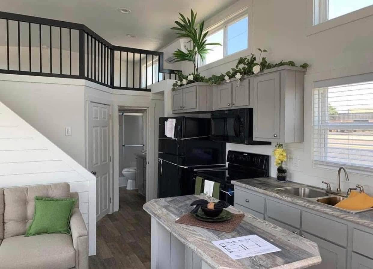 A modern tiny home interior with a cozy kitchen featuring gray cabinets, black appliances, and a small island. Plants add greenery, and a loft area with a black railing overlooks the space. Light streams through multiple windows, creating a bright and inviting atmosphere.