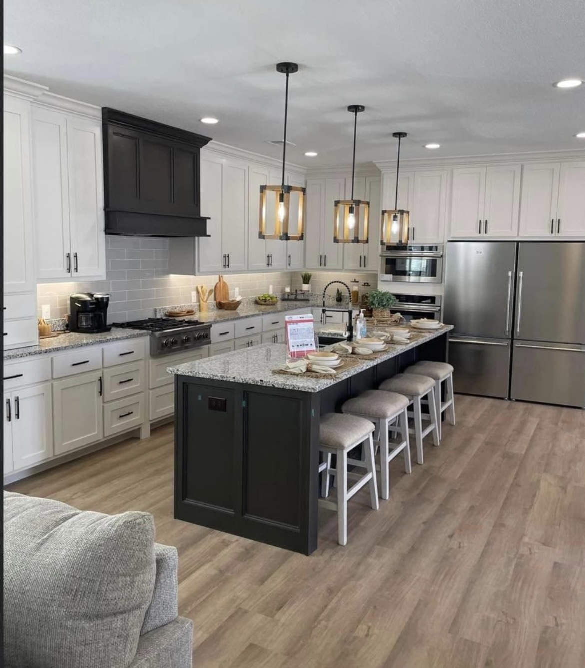 Modern kitchen with a large island, granite countertops, and four pendant lights. White cabinets, stainless steel appliances, and wood flooring create a warm ambiance.