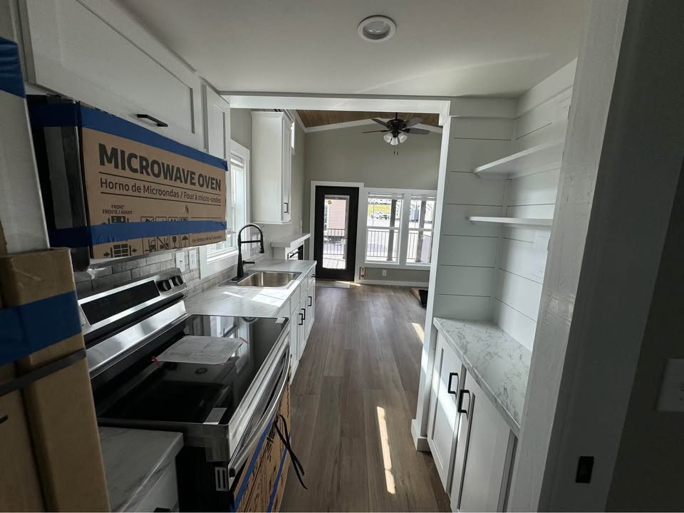 Modern, narrow kitchen with white cabinetry, stainless steel appliances, and wood flooring. Sunlit, cozy dining area visible through door.