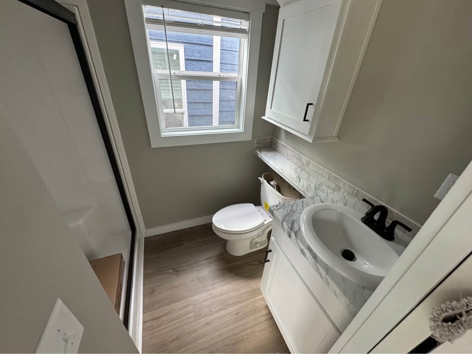 Small bathroom with gray walls, wood floor, and a window. Features a shower, white sink with marble countertop, and a toilet. Compact and tidy space.