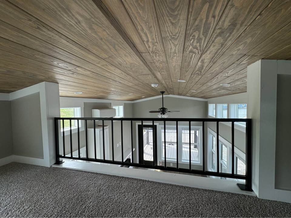 Loft with wood-paneled ceiling and black railing overlooks large windows and a ceiling fan. The space feels airy and bright, with neutral tones.