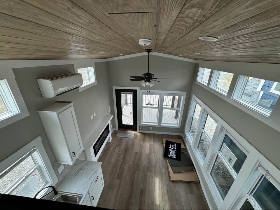 View of a modern tiny house interior with large windows, wood floors, and ceiling. Features include a ceiling fan, white cabinets, and a black door.