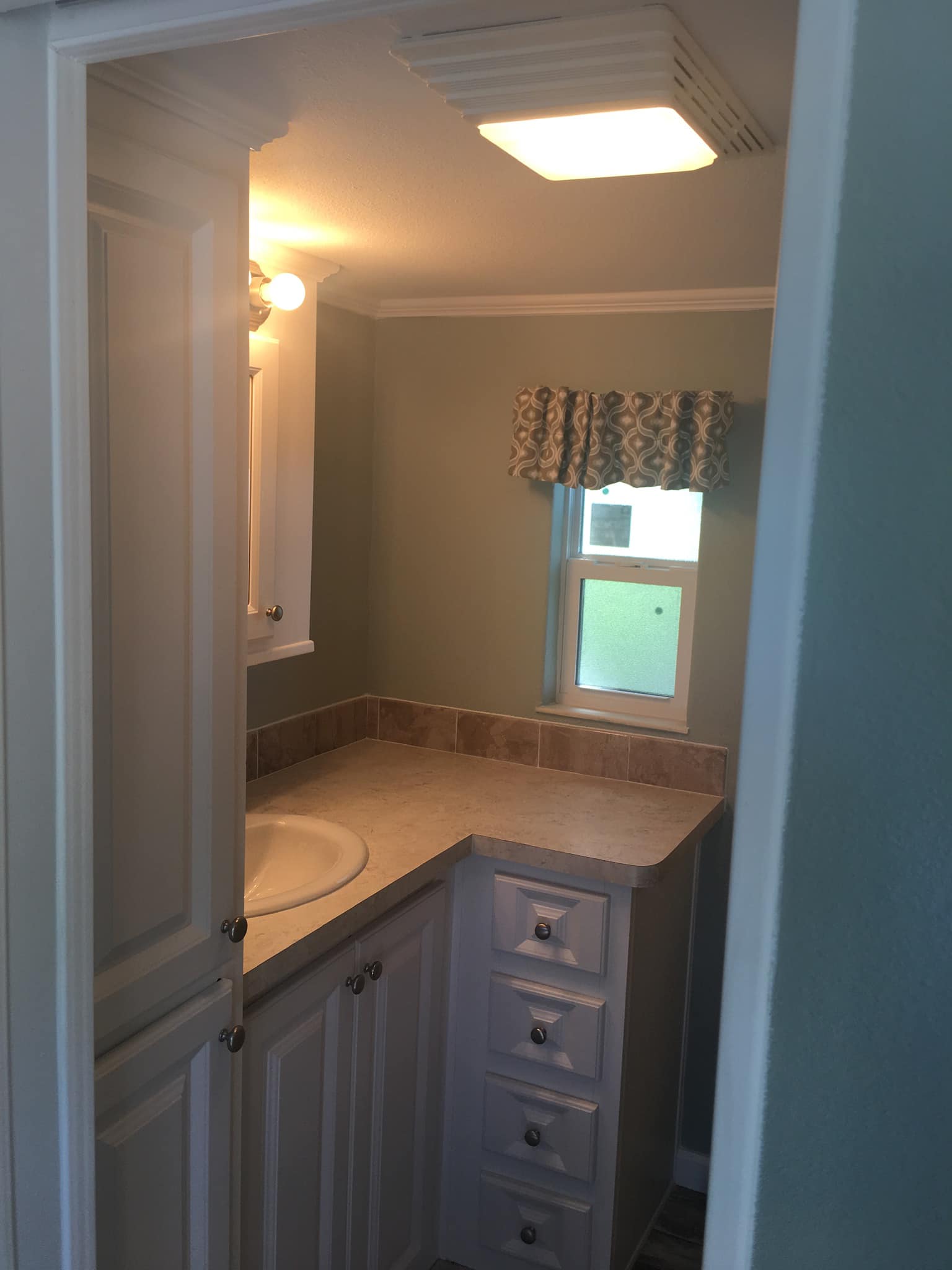 Compact bathroom with light green walls, featuring a small window with patterned curtains. There’s a white cabinet, sink, and a tan countertop.