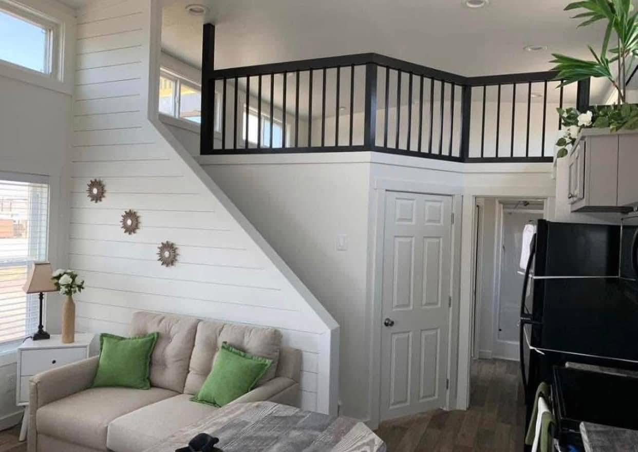 Cozy living room with light sofa and green pillows. White shiplap wall with decorative gears. Loft area above, black railing. Bright, welcoming space.
