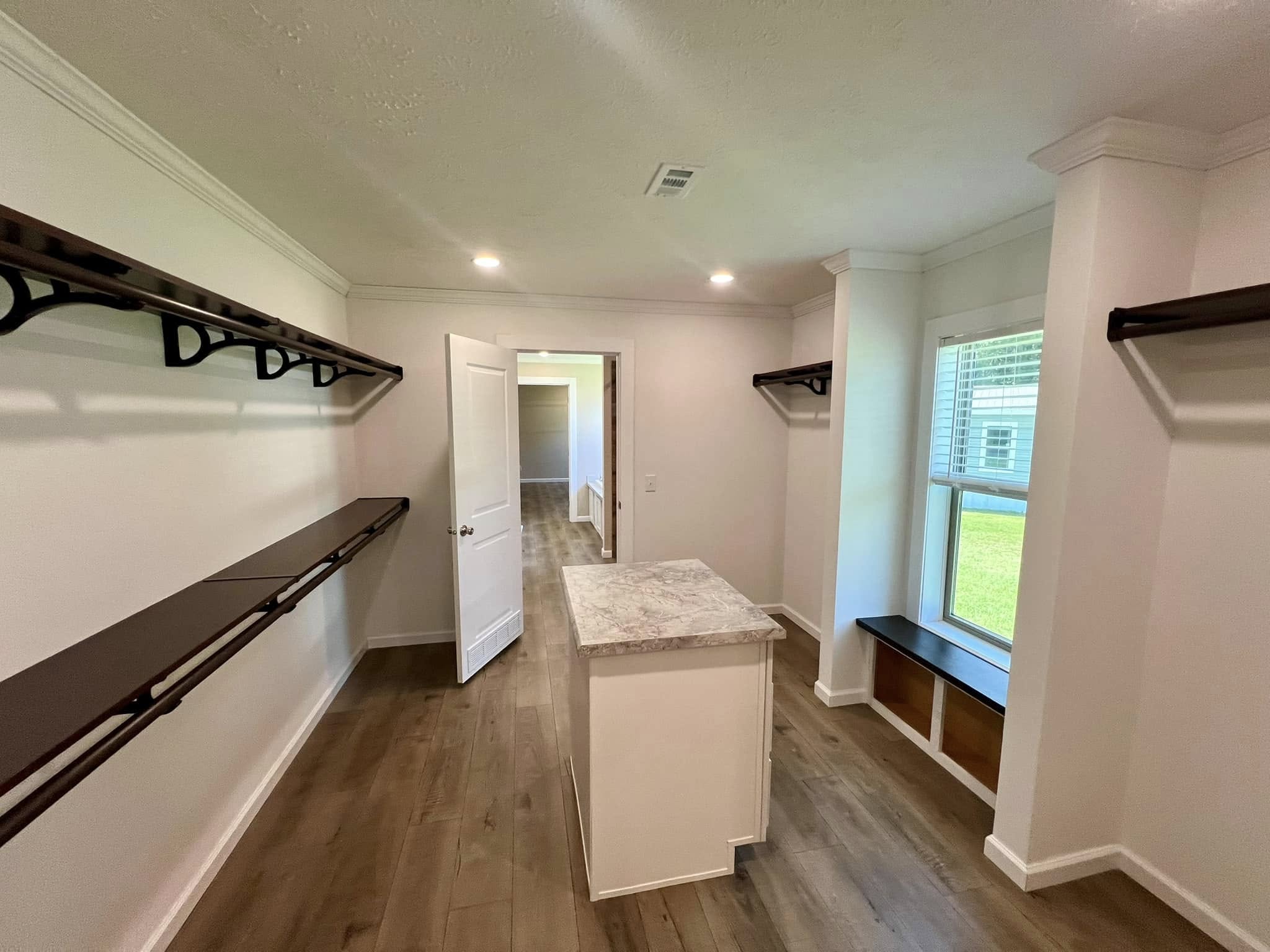 Spacious walk-in closet with wooden shelves and a central island, featuring a bright window nook and light wood flooring, conveying a modern and organized feel.