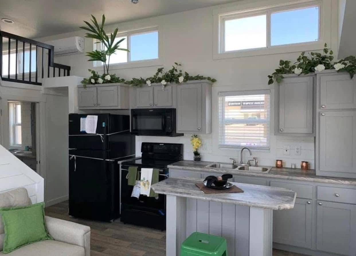Bright kitchen with gray cabinets, marble countertops, and black appliances. Greenery decorates the top cabinets. A cozy loft is visible above.