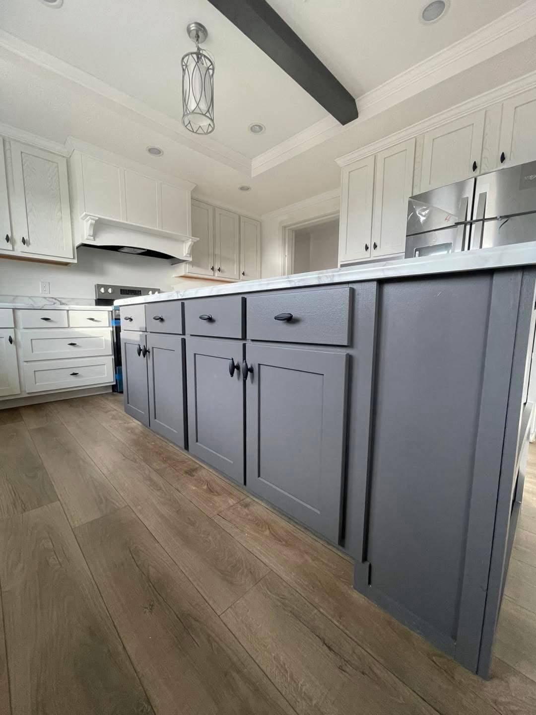 Modern kitchen featuring a large gray island with black handles, white cabinets, light wood flooring, and a decorative light fixture above.
