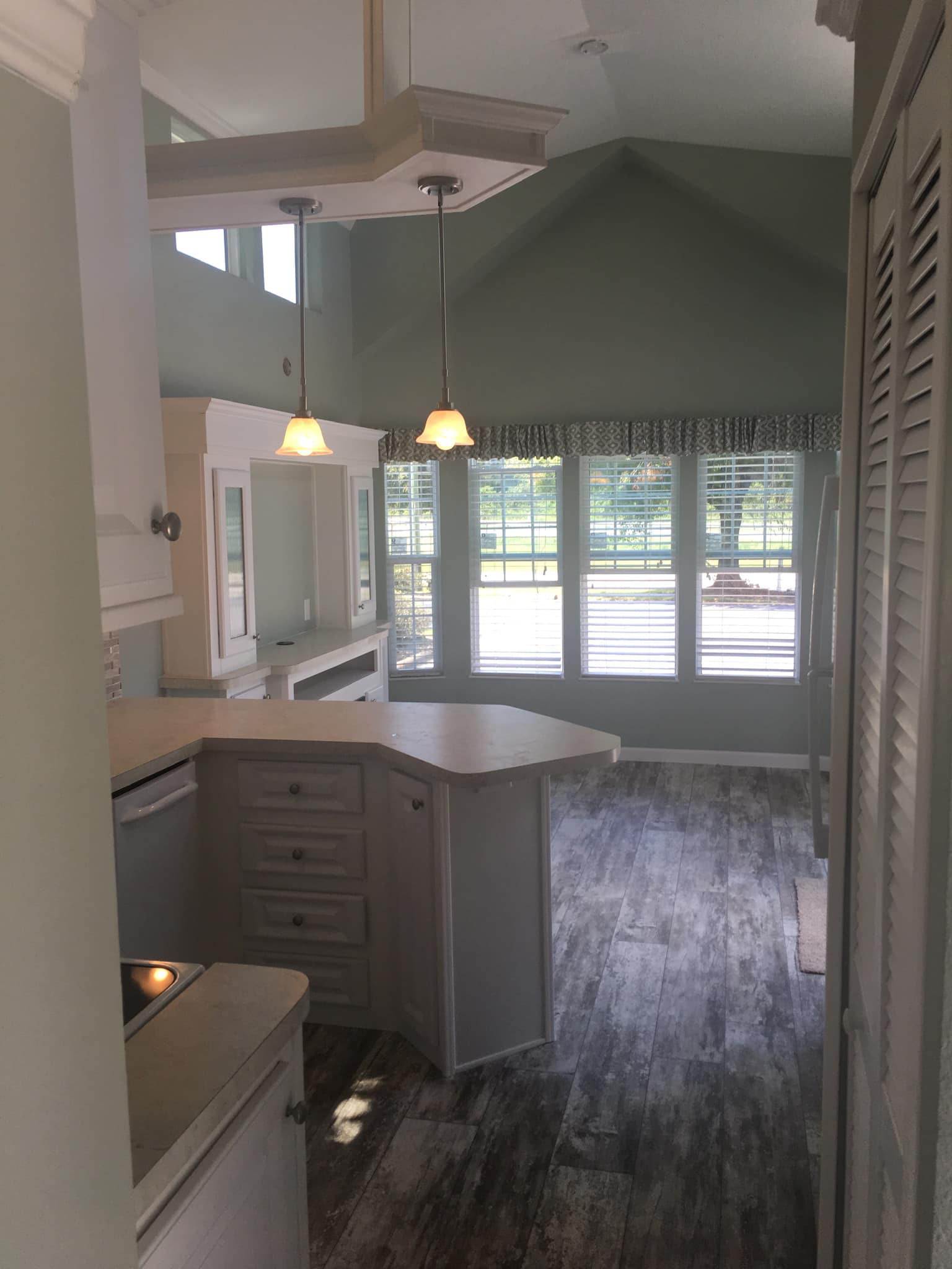 Bright kitchen with high ceilings, pendant lights, and large windows letting in natural light. Gray wood-look floor and light-colored cabinets. Cozy and airy.