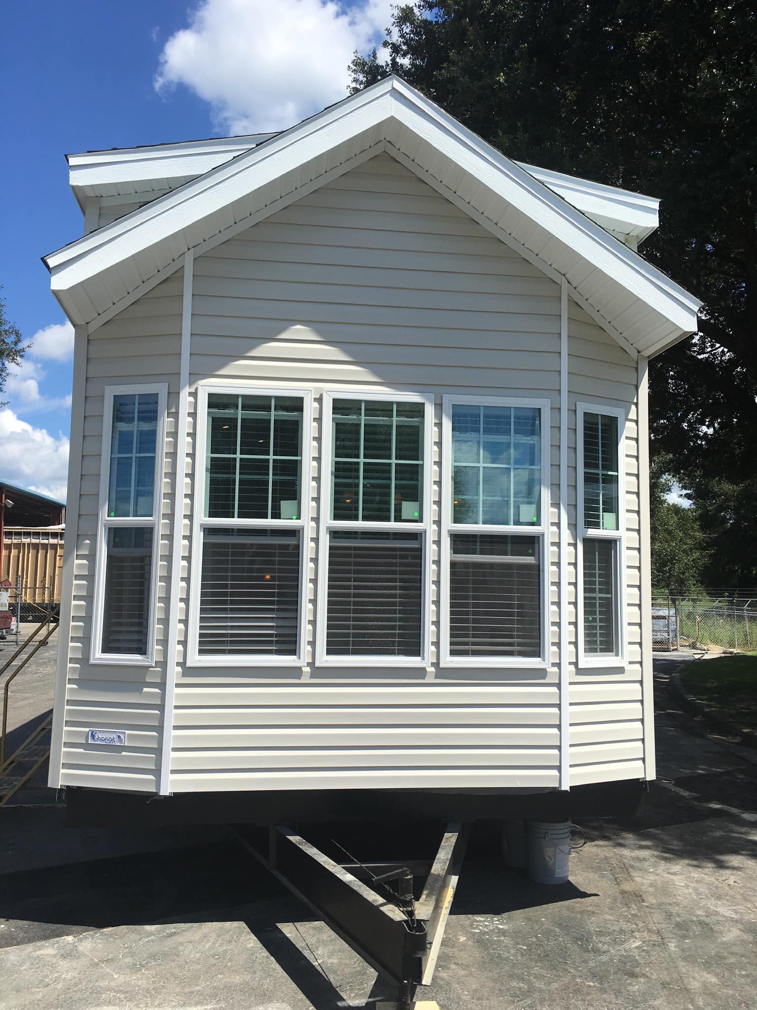 A small, beige mobile home with a peaked roof and large windows sits on a sunny lot. Shadows and bright sky convey a peaceful, sunny day.