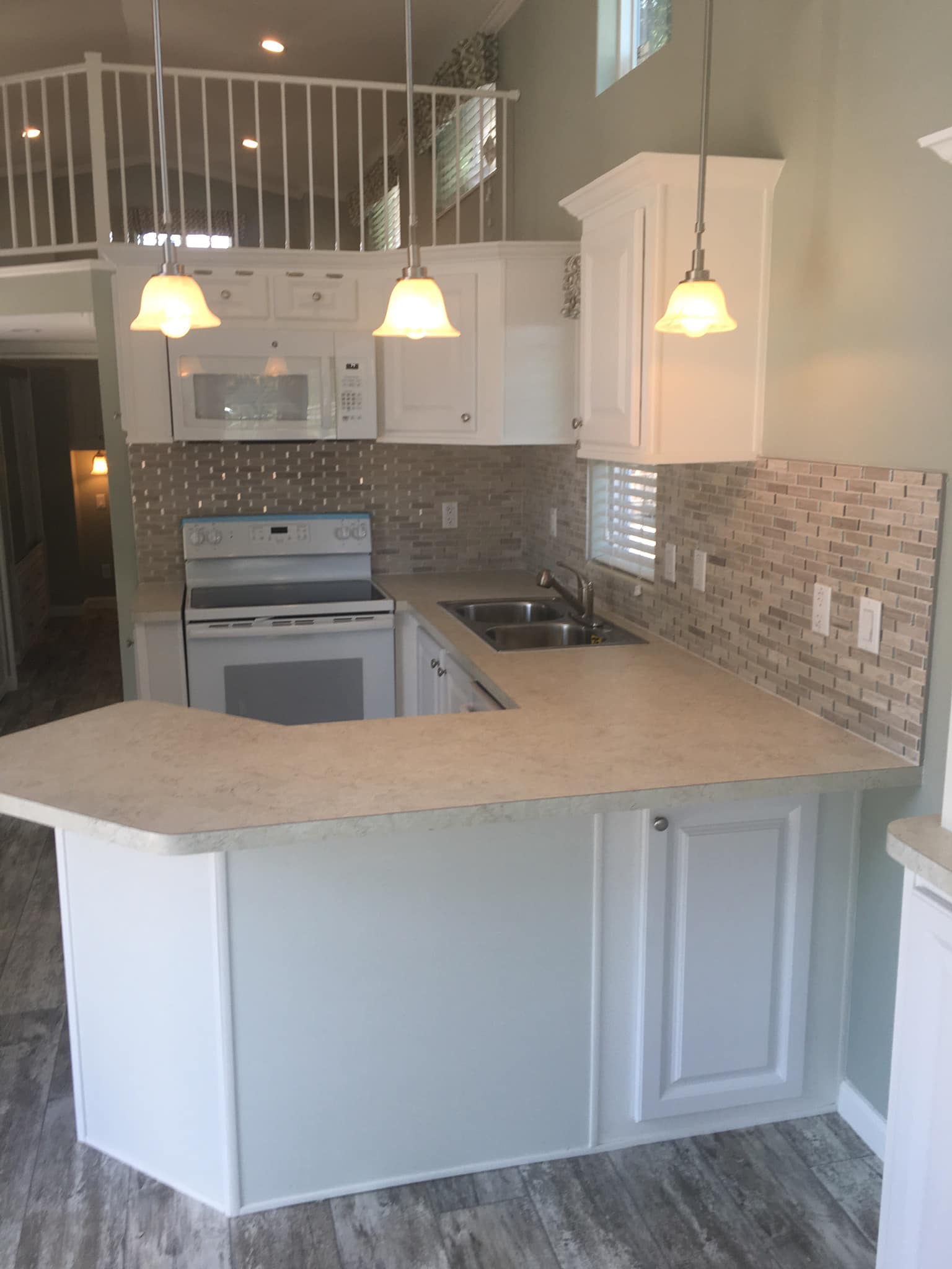 Compact kitchen with white cabinets, light countertops, and mosaic backsplash. Includes a stove, microwave, and double sink. Warm lighting from pendant lamps.