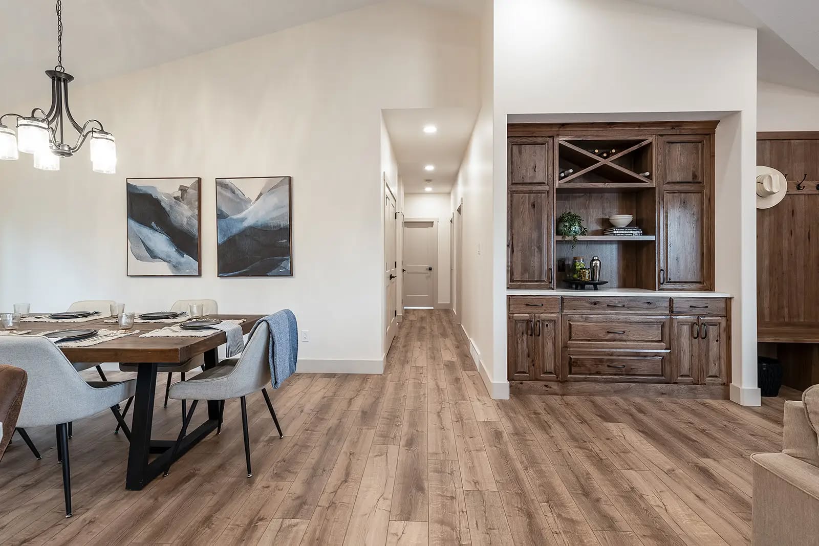 Modern dining room with wood flooring, featuring a wooden table set for four with gray chairs. Left wall has two abstract paintings; right side has a wooden built-in cabinet. A hanging chandelier adds elegance. Neutral tones create a warm ambiance.