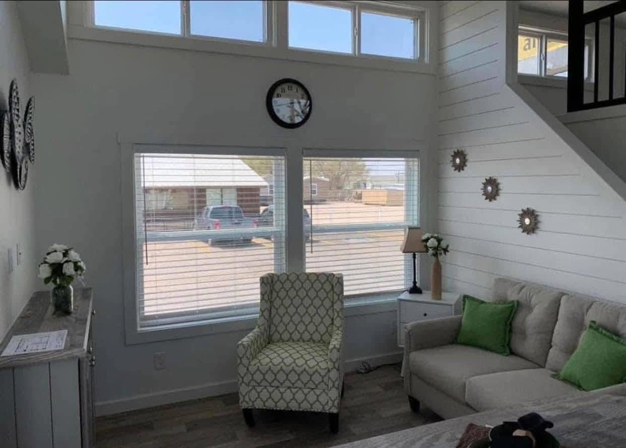 Bright room with large windows, patterned armchair, and beige couch with green pillows. Wall clock and decorative gears add charm. Cozy atmosphere.