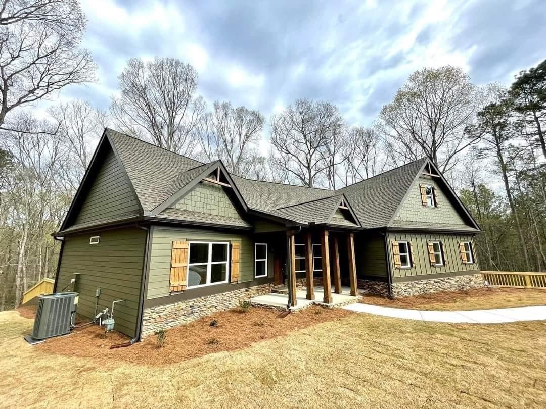 Modern cabin-style house with green siding, wooden shutters, and stone accents. Set amid bare trees under a cloudy sky, it feels serene and natural.