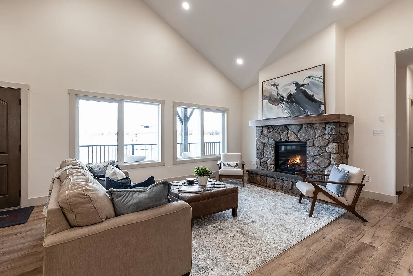 Spacious living room with vaulted ceiling, stone fireplace, and large windows. Earth-toned decor includes a beige sofa and wooden furniture, creating a cozy ambiance.