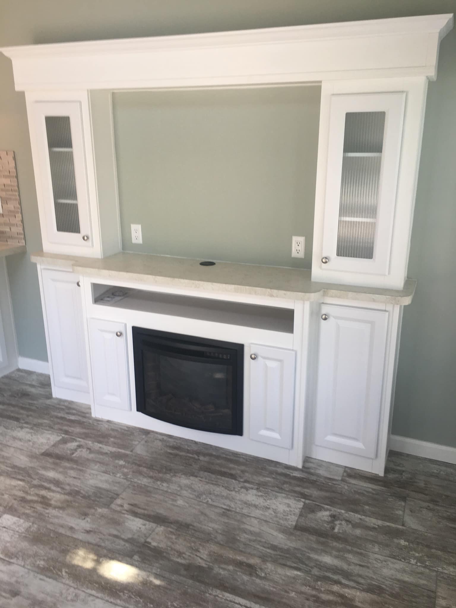 White built-in cabinet with a fireplace at the center, flanked by two glass-paneled doors, set against a light gray wall on wood-patterned flooring.