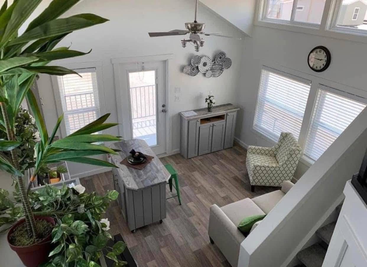 A cozy living room with wooden flooring, white walls, and large windows allowing natural light. Features a sofa, armchair, greenery, and wall clock.