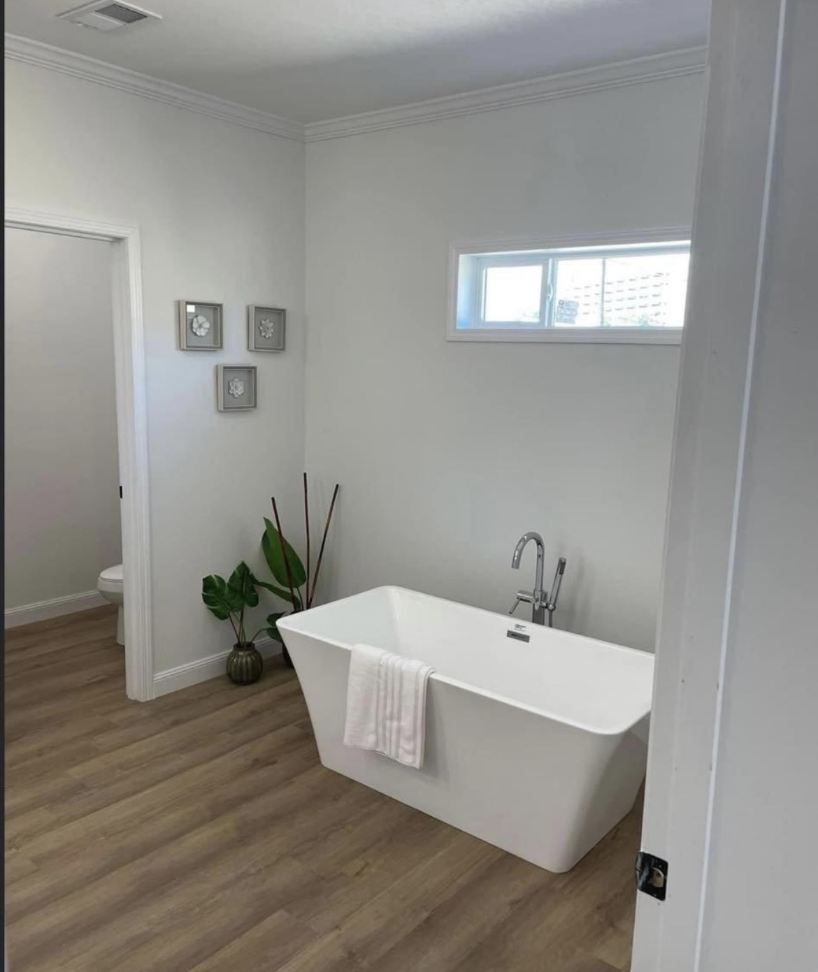 Minimalist bathroom with a freestanding white bathtub on light wood flooring. A window above, decorative plants, and framed art create a serene atmosphere.