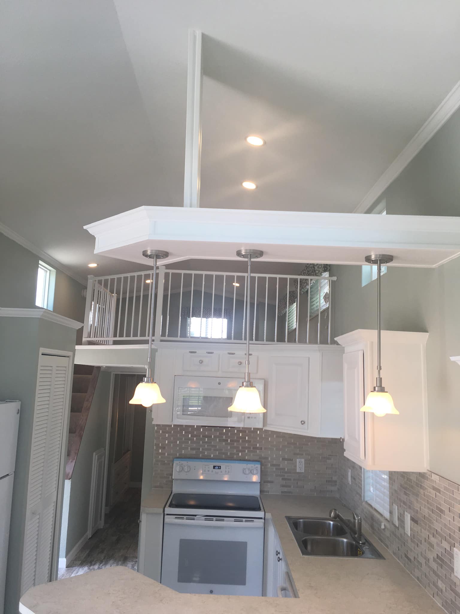 Modern tiny house kitchen with high ceiling. Features white cabinets, stainless steel appliances, loft railings above, and three hanging pendant lights. Cozy and bright.