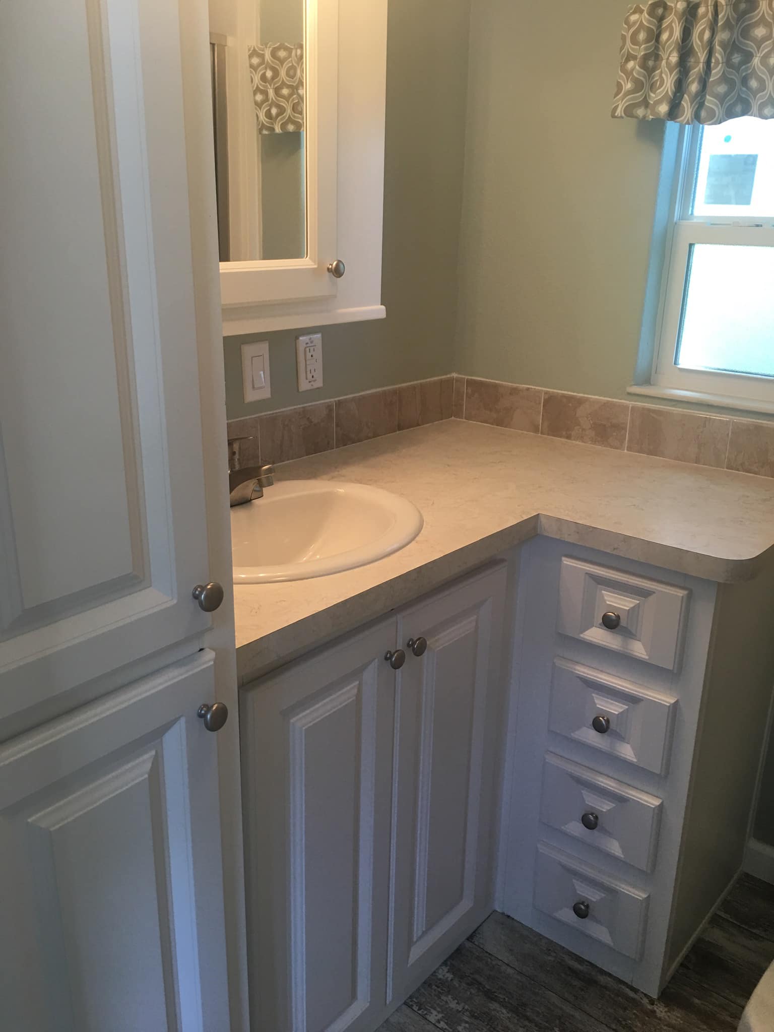 Compact bathroom with light green walls, featuring white cabinets and a beige countertop with a round sink. A small window is adorned with floral curtains.