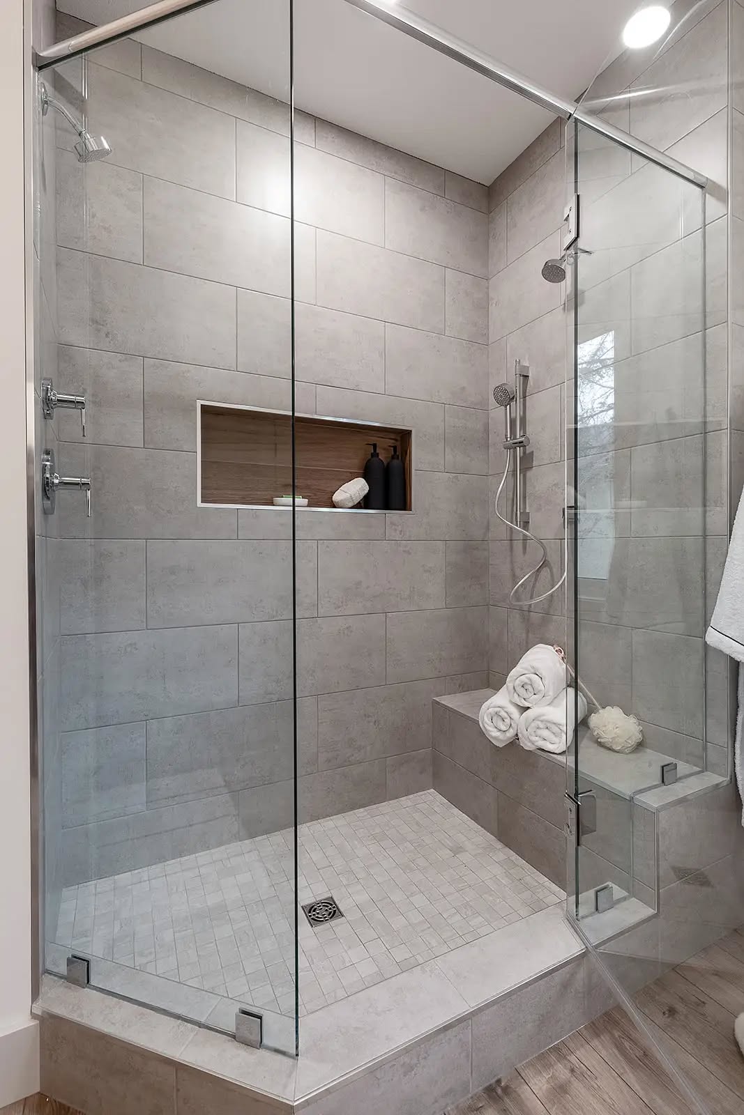 Modern shower with glass enclosure, gray tiles, and built-in shelf holding toiletries. A bench with rolled towels adds a spa-like feel.