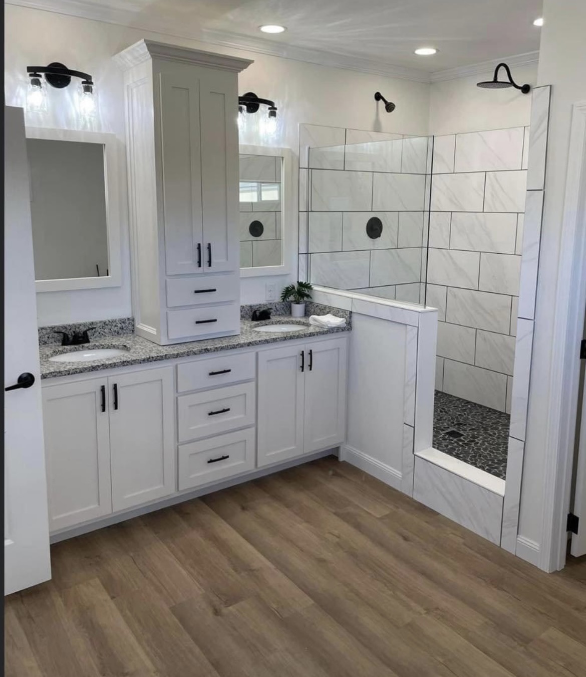 Modern bathroom with white cabinets, granite countertops, and dual mirrors. Features a walk-in shower with large tiles and wood-style flooring.