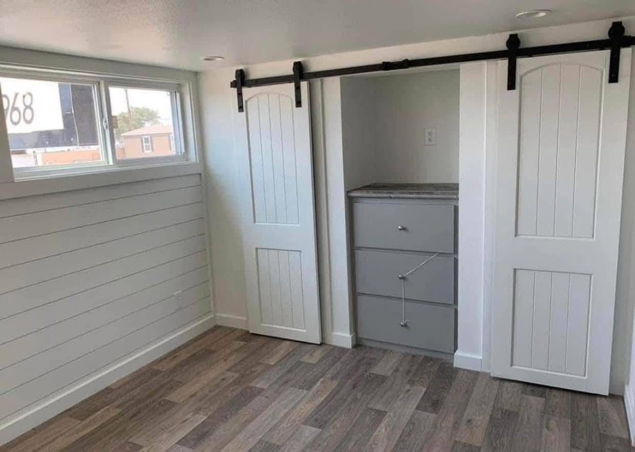 Compact room with gray wood flooring and white walls. Features a small closet with sliding barn doors, housing a gray dresser with three drawers. Bright and minimalist.